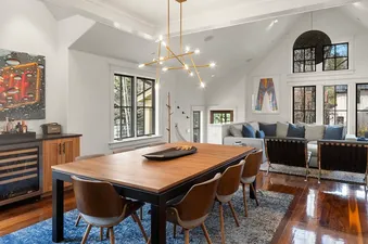 An open-concept dining and living area with a long wooden table, modern chairs, a gold chandelier, and large windows offering views of the outdoors.