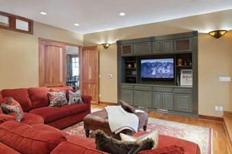 A cozy entertainment area with a red sectional sofa, a leather ottoman, and a built-in olive green media cabinet with a TV displaying a mountain scene.