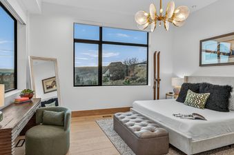 A luxurious bedroom at Nocturne View with a king-sized bed, tufted ottoman, stylish desk, accent chair, and large windows showcasing mountain views.