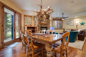 A rustic-chic dining area with an antler chandelier and a wooden table with a glass top, leading into a cozy living space with a stone fireplace, visible through a large glass door.