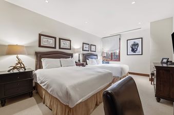 A luxurious bedroom featuring two queen beds with brown leather headboards, elegant bedside tables with lamps, framed artwork, and a large window with a white Roman shade.