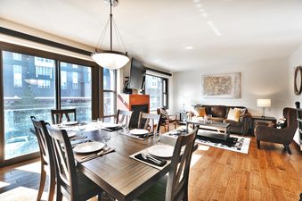 A spacious, open-concept living and dining area in Lowell 303, featuring a large dining table with eight chairs, a comfortable brown leather sofa, an armchair, a fireplace, and large windows.