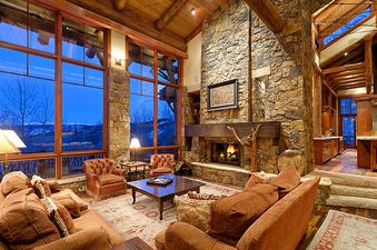 The living room of the Treehouse on West Fork features a large stone fireplace, comfortable brown seating, and expansive windows showcasing an exterior view at twilight.