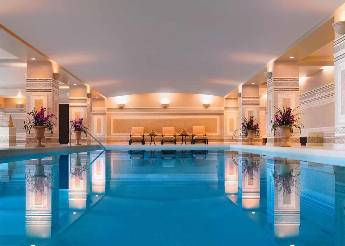 An expansive indoor pool gleams with inviting blue water, bordered by elegant decorative pillars and soft, ambient lighting, with comfortable chairs arranged along the far wall.