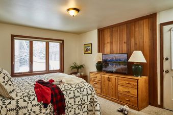 Cozy bedroom at Teewinot B3 featuring a patterned bed quilt, wooden dresser with TV, and a window displaying a snowy winter view.