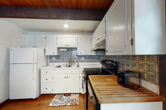 A fully equipped kitchen with white cabinets, a double stainless steel sink, a black electric stove, a white refrigerator, and a vibrant blue and yellow tiled backsplash.