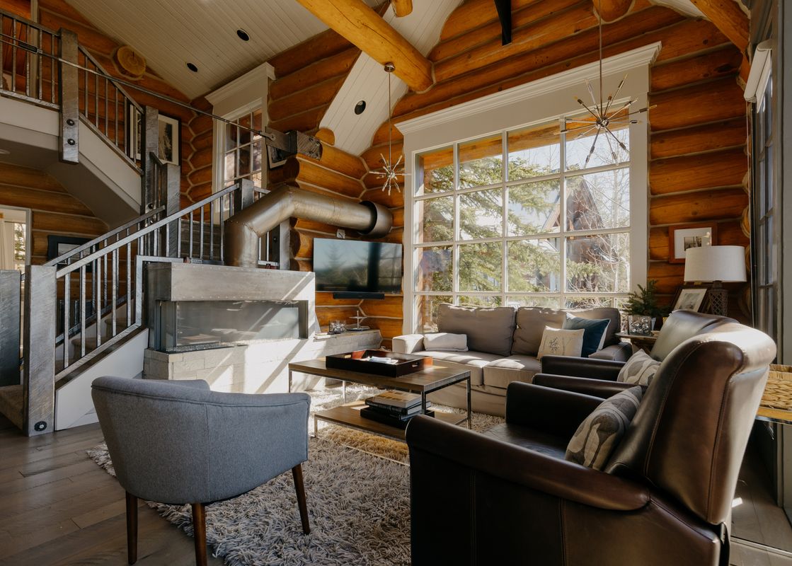 The living room of the Summit Cabin at See Forever features a large fireplace, a sofa, two armchairs, and expansive windows offering views of the outdoors.