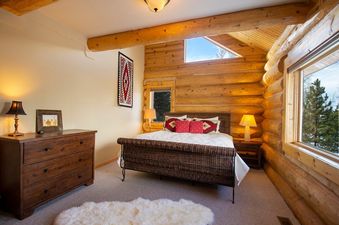 A cozy bedroom in Miles Behind with a wooden dresser, a woven bed, and large windows revealing a natural outdoor view.