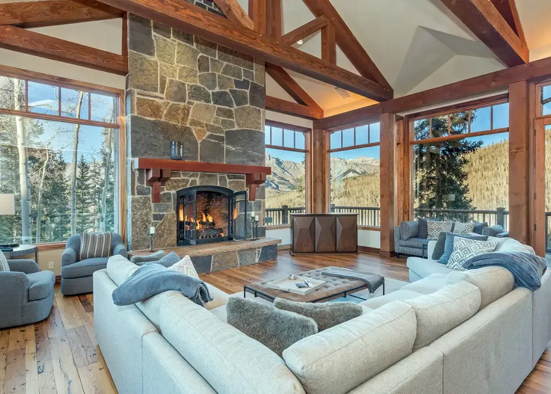 The living room in Two Trails features a large stone fireplace with a roaring fire, comfortable seating, and expansive windows that offer breathtaking mountain views.