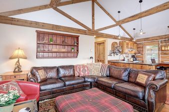 The living room of Moonlight Basin 9 Cabin Hollow features a large leather sectional sofa, a red armchair, a plaid ottoman, and a wooden coffee table. The room also has a large window with a view of the forest.