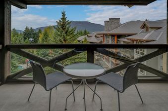 A private balcony offers an intimate setting with a small round table and two chairs, ideal for enjoying the mountain and forest views, along with glimpses of nearby buildings.