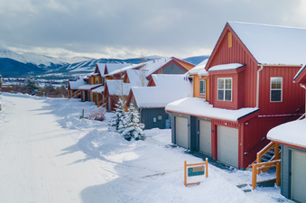 A row of charming townhomes covered in fresh snow, with a snowy street in front and majestic snow-capped mountains in the background under a cloudy sky.