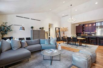 A modern living room with a sectional sofa, a large rug, a coffee table, two poufs, and a kitchen in the background.