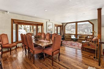 The dining area and living space at Woodrun V Residence 5 feature a large wooden dining table with upholstered chairs, a cozy living area with armchairs and a decorative chest, and expansive windows offering snowy mountain views.