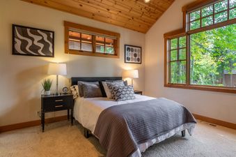 A cozy bedroom in the Northstar Sierra Gold Home with a bed, nightstands, lamps, and large windows looking out onto green trees.