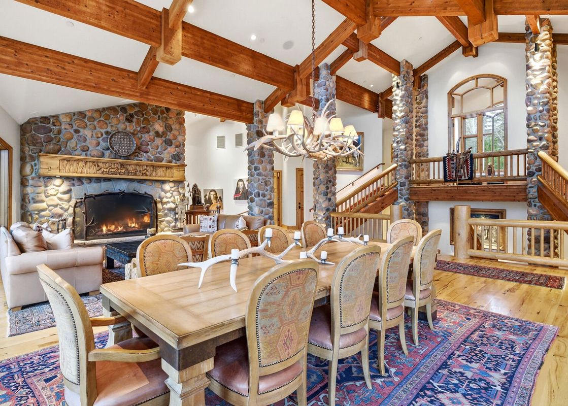 A rustic dining room features a large wooden table with an antler candelabra, surrounded by upholstered chairs, with a stone fireplace and grand staircase in the background.