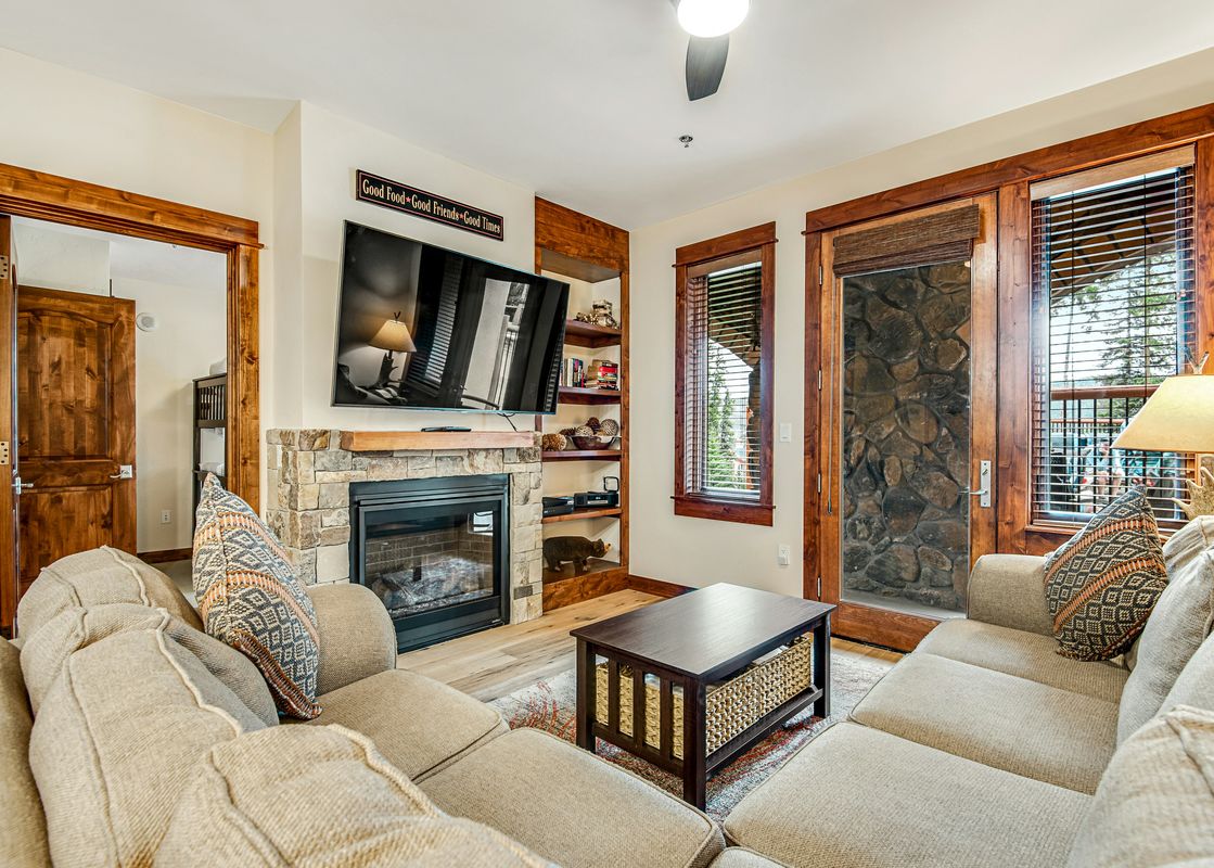 A cozy living room at BlueSky Breckenridge 407 featuring a stone fireplace, television, comfortable sofas, and wooden accents, with outdoor views through the glass doors.