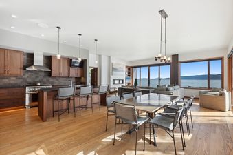 An expansive open-concept living area at the Swan Lake House, featuring a modern kitchen with wooden cabinetry, a dining area with a large table, and a lounge with a fireplace, all overlooking a stunning lake and mountain view through large windows.