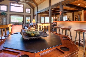 A wide-angle shot of the Antler Ridge Lodge's main living area, featuring a large wooden poker table in the foreground, a cozy living room with a fireplace, and a bar area with stools.