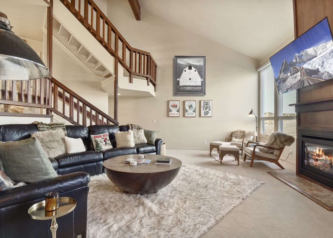 A bright living room features a dark leather sectional, a large round wooden coffee table on a fluffy area rug, and a fireplace with a mounted television displaying a snowy mountain scene.