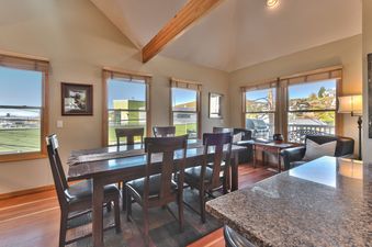 A spacious dining area with a long wooden table and chairs, surrounded by large windows offering scenic views, a lounge area, and a kitchen counter with a granite top.