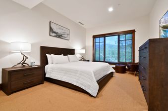 A luxurious king bedroom at Arrowleaf Lodge 107, featuring a king-sized bed, dark wooden furniture, a cozy armchair, and a large window offering a view of the outdoors.