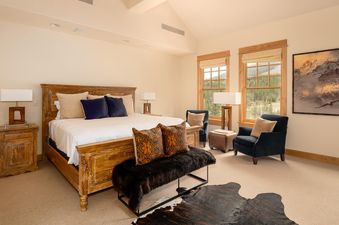 The master bedroom of Moonlight Basin Penthouse 3 features a king-size bed with plush bedding, two nightstands, and two chairs near the windows that offer stunning views of the mountains.