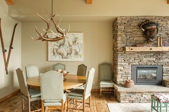 A rustic dining area with a round table and an antler chandelier, adjacent to a stone fireplace.