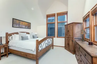 A serene bedroom in the South Pacific Townhouse with a large wooden bed, white bedding, panoramic forest artwork, and rustic wooden furniture, all under vaulted ceilings with natural light from multiple windows.