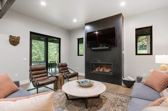 A modern living room with a gray sofa, two brown leather armchairs, a round coffee table on a patterned rug, and a black fireplace feature wall with an integrated television.