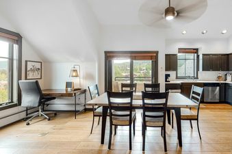An open-concept living area in Main Street Station 3405, featuring a dining table, a workspace with a desk and chair, and a kitchen with dark cabinetry and stainless steel appliances.