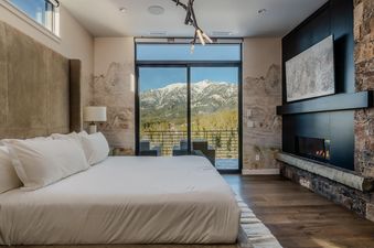The master bedroom of Moonlight Basin 7 Speedwell Drive offers a stunning view of the mountains from a large window. The bedroom is decorated with a neutral color palette and features a plush bed with white linens.  A fireplace is visible on the right sid
