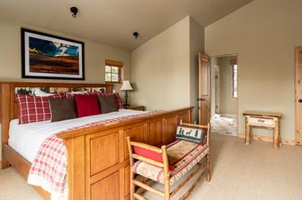 A king-sized bed with a wooden headboard and a red plaid blanket sits in the center of the room, with a wooden chair beside it. The room is decorated with a western-style theme and there is an open doorway to the bathroom on the right.