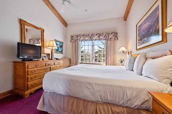 The primary bedroom in The Charter E2205 features a king bed with white bedding, a television on a wooden dresser, a window with a floral valance and drapes, and a purple carpet.