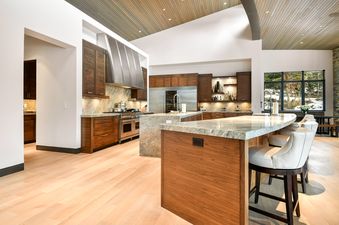 A modern kitchen with dark wood cabinets, light granite countertops, a large stainless steel range hood, and two kitchen islands, one with seating, all set on light wood floors.