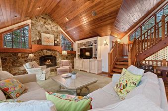 The Lake View Escape living room showcases a large stone fireplace, wood-paneled vaulted ceilings, and comfortable seating, with large windows offering a view of the outdoors.