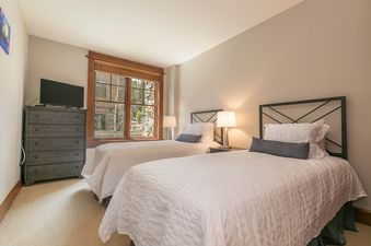 Two twin beds in a bedroom, with a dresser, a flat screen TV, a window, and lamps