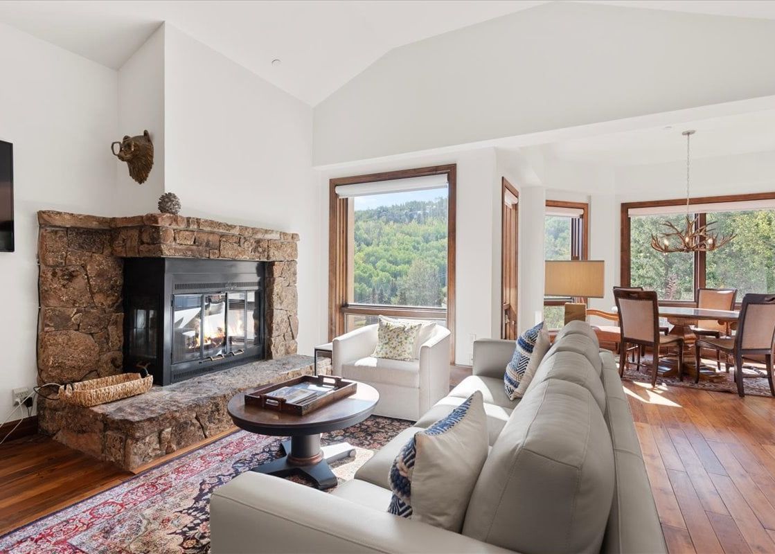 A bright, open-concept living area at Chamonix 50 featuring a stone fireplace, comfortable white leather seating, and large windows with scenic forest views.