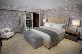 The Clubhouse, a spacious bedroom with a king-size bed, a plush grey ottoman, a comfortable chair, and a view to the outside.