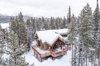 A snowy chalet-style home with a large deck and hot tub, surrounded by snow-covered pine trees and distant mountains under a gray sky.
