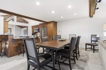 A spacious dining area featuring a large wooden table with black leather chairs, connected to a modern kitchen with wooden cabinets and stainless steel appliances.