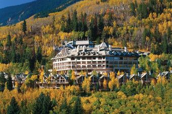 The Pines Lodge stands majestically amidst a vibrant autumn landscape, surrounded by golden-leaved trees and smaller luxury homes, under a clear blue sky.