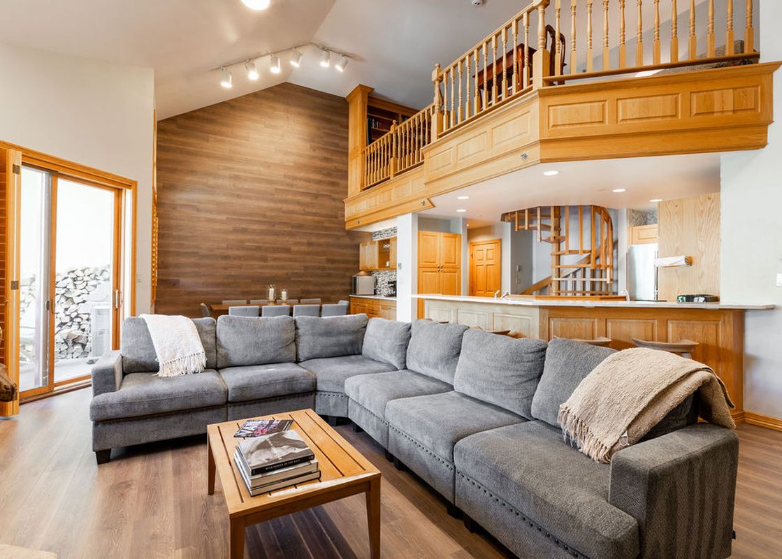 A spacious living area with a large gray sectional sofa and a wooden coffee table, featuring a high wooden accent wall and an upper-level loft area with a wooden railing.