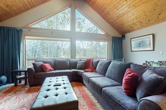 A spacious living room featuring a large gray sectional sofa, a black tufted ottoman, and expansive windows offering views of an outdoor forest.