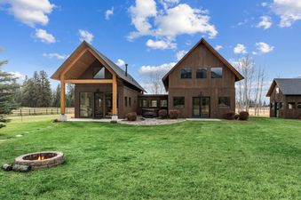 A modern lodge with dark brown siding and gabled roofs, featuring a lush green lawn, a stone fire pit, and a hot tub.