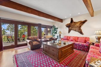 A rustic yet elegant living room with red sofas, leather armchairs, a large wooden coffee table, and a bear skin rug on the wall, offering stunning views of pine trees and a lake from a balcony.