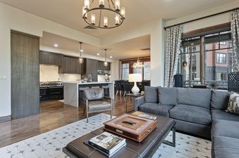 A spacious open-concept living area with a grey sectional sofa, brown leather ottoman, and a modern kitchen with dark wood cabinets and marble countertops.