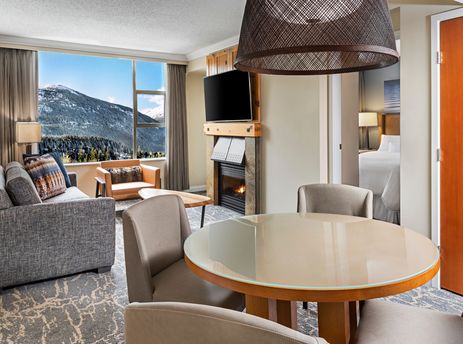 The Westin Resort One Bedroom Mountain View offers a cozy living area with a fireplace, comfortable seating, and a dining table, all set against a stunning mountain backdrop visible through large windows.