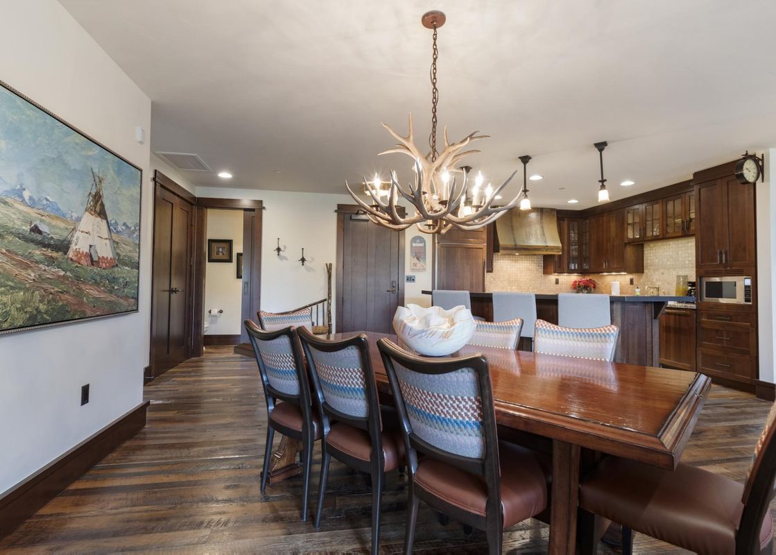 A dining table with chairs, lit by an antler chandelier, with a kitchen visible in the background.