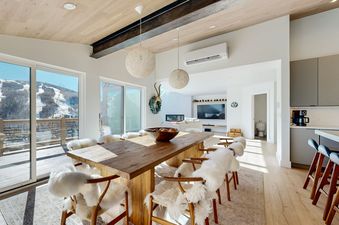 A spacious dining area in Luxe Lookout with a large wooden table and chairs draped in white sheepskin, overlooking snow-covered mountains through expansive glass doors.